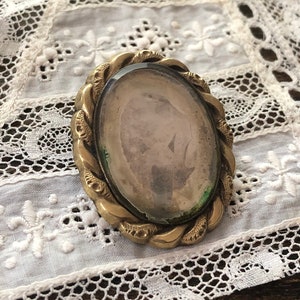 May include: A gold-toned oval locket with a glass front and a braided design around the edge. The locket is on a white lace background.