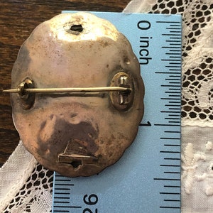 May include: A silver-toned brooch with a round, scalloped edge. The brooch has a pin and catch on the back. The brooch is about 1 inch in diameter.