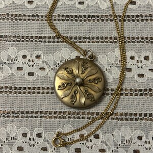 Puede incluir: Un collar de oro con un colgante redondo que presenta un dise&ntilde;o floral y una perla en el centro.