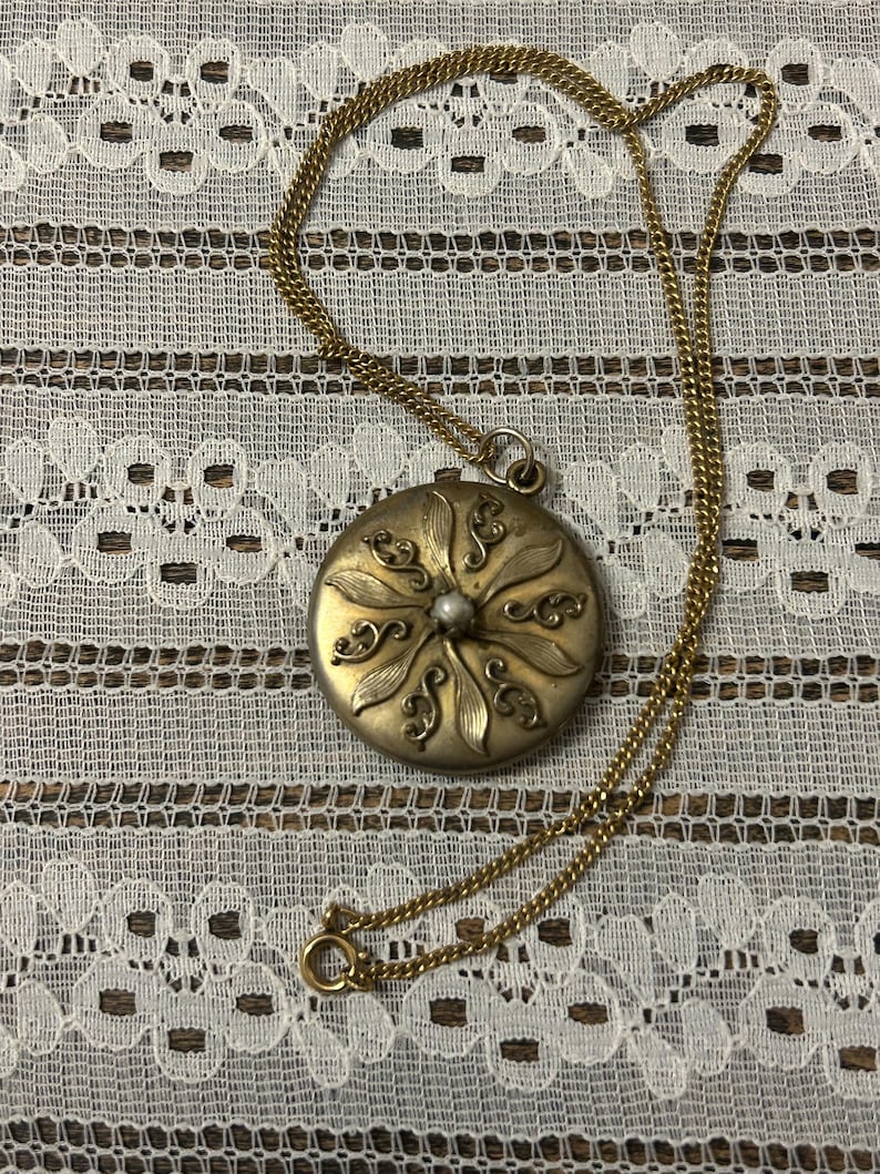 Puede incluir: Un collar de oro con un colgante redondo que presenta un dise&ntilde;o floral y una perla en el centro.