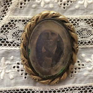 May include: A vintage oval-shaped locket with a gold-toned metal frame and a glass cover. The locket features a detailed design of a braided rope around the edge. Inside the locket is a faded black and white photograph of a person.
