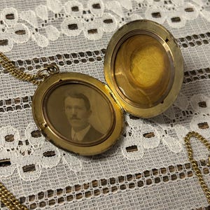 Puede incluir: Un collar de oro con un medall&oacute;n y una cadena. El medall&oacute;n est&aacute; abierto, revelando una foto en blanco y negro de un hombre con bigote. El medall&oacute;n tiene una superficie lisa y brillante.