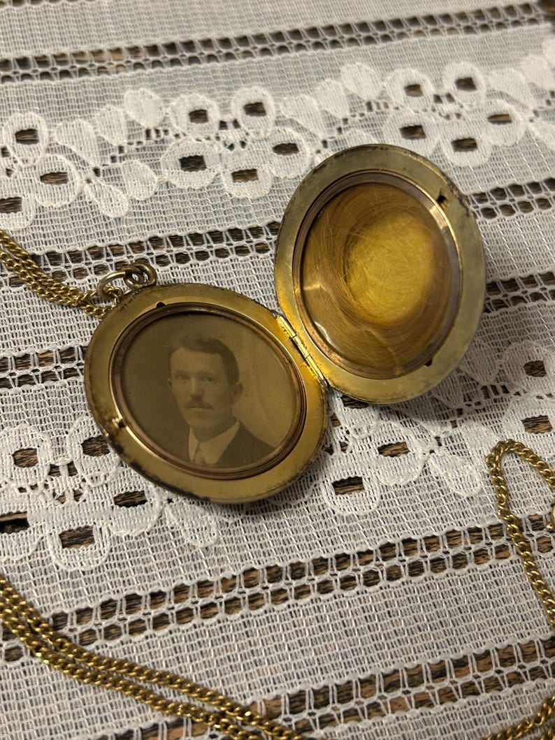 Puede incluir: Un collar de oro con un medall&oacute;n y una cadena. El medall&oacute;n est&aacute; abierto, revelando una foto en blanco y negro de un hombre con bigote. El medall&oacute;n tiene una superficie lisa y brillante.