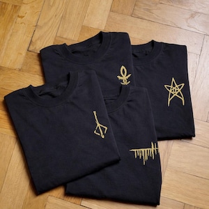 May include: Four black t-shirts with gold-colored embroidered designs. Each shirt features a different symbol: an ankh, a star, a stylized weapon, and a series of vertical lines. The shirts are neatly folded on a wooden surface.