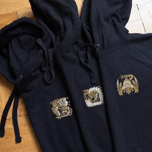 May include: Three navy blue hoodies with gold dragon designs. Each has a drawstring hood and a kangaroo pocket. The dragon designs vary in style and are embroidered on the front of the sweatshirts.