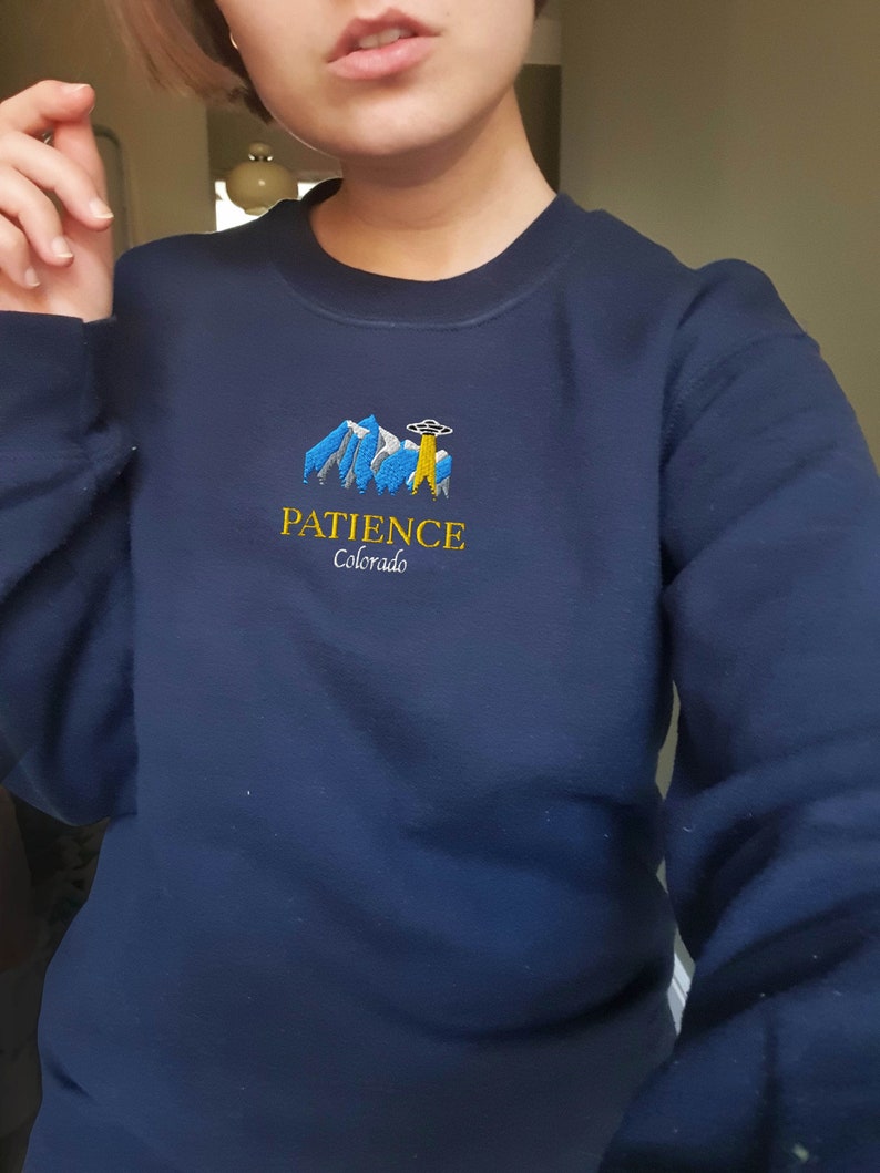 May include: A navy blue sweatshirt with an embroidered design of mountains, a UFO, and the text "PATIENCE Colorado".
