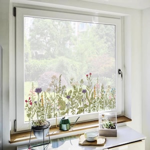 Könnte beinhalten: Ein Fenster mit floralem Design, das Wildblumen in Lila, Rot, Gelb und Grün zeigt. Das Fenster ist in einem weißen Rahmen eingefasst. Ein kleiner Blumentopf und eine Gießkanne stehen auf der Fensterbank, mit Blick auf einen Garten.