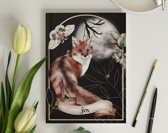 Op de afbeelding: Een zwart notitieboek met een gouden frame en een aquarel-illustratie van een vos in een bosrijke omgeving. De vos is roodbruin met een witte borst en een borstelige staart. De achtergrond is donker met een volle maan en een nachtvlinder. De tekst "Fox" is geschreven in gouden letters op een witte banner.