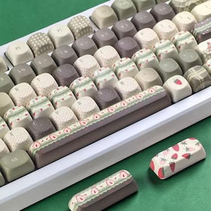 May include: A white mechanical keyboard with custom keycaps. The keycaps feature various designs, including strawberries, polka dots, and patterned stripes in shades of pink, green, and brown. The keyboard has a white frame and a green background.