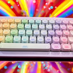 May include: A pastel-colored keyboard with cat-shaped keycaps. The keycaps are arranged in a standard layout, with each keycap featuring a cat face design. The keyboard has a white space bar and a colorful background.