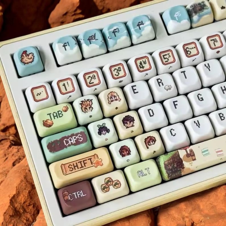 Op de afbeelding: Een aangepast mechanisch toetsenbord met witte en beige toetsen. De toetsen hebben verschillende kleurrijke ontwerpen, waaronder gepixelde karakters, wolken en symbolen. De toetsen "TAB", "CAPS", "SHIFT" en "CTRL" zijn gelabeld. Het toetsenbord heeft een lichtgekleurde lijst.