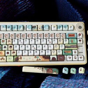Op de afbeelding: Een cr&egrave;me mechanisch toetsenbord met aangepaste keycaps. De keycaps bevatten illustraties en tekst, waaronder cijfers, letters en symbolen. Het toetsenbord heeft een gele rand en een bijpassende polssteun. Extra keycaps liggen verspreid.