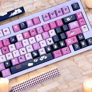 Può includere: Una tastiera meccanica personalizzata con una cornice viola chiaro e tasti nei toni del rosa, nero e bianco. I tasti presentano vari simboli e illustrazioni, tra cui cuori, conigli e semi di carte da gioco. Tasti aggiuntivi sono posizionati di lato.