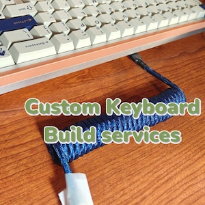 May include: A custom keyboard with a white and beige keycap set, a blue and black coiled cable, and the text "Custom Keyboard Build Services" on a wooden surface.