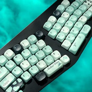 Può includere: Una tastiera meccanica nera con tasti personalizzati. I tasti sono di colore verde acqua chiaro con lettere e simboli bianchi. Alcuni tasti hanno disegni unici, tra cui una barca a vela e le parole "Mulled Wine".