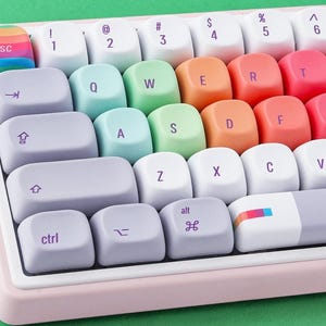 May include: Close-up of a mechanical keyboard with pastel-colored keycaps. The keys are arranged in a standard layout, with a rainbow-colored escape key. The keyboard has a light pink frame and sits on a green surface.