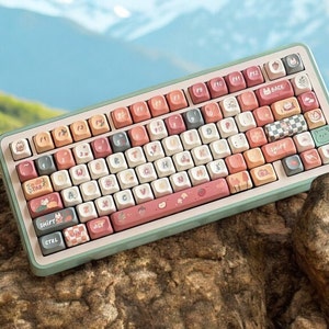 Può includere: Una tastiera meccanica con tasti personalizzati dai colori pastello. I tasti presentano varie illustrazioni, tra cui animali, piante e motivi. La tastiera ha una cornice verde chiaro ed è ambientata su uno sfondo naturale di rocce e un paesaggio montano.