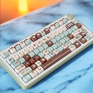Può includere: Una tastiera meccanica verde menta e bianca con tasti personalizzati. I tasti presentano disegni di barrette di cioccolato, cuori e illustrazioni. La tastiera ha un design a impronta di zampa nell'angolo in alto a destra.