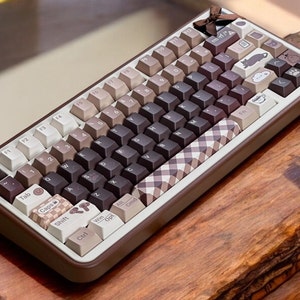 May include: A mechanical keyboard in shades of brown and beige. The keycaps are decorated with patterns and illustrations, including a gingham design, cats, and a coffee cup. The keyboard has a brown frame and a small bow at the top.