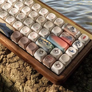Puede incluir: Un teclado de madera con teclas blancas y teclas personalizadas con diseños de inspiración japonesa, que incluyen una montaña, una pagoda y nubes. El teclado está apoyado sobre una superficie rocosa marrón.