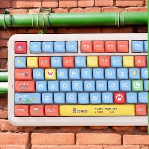 Può includere: Una tastiera bianca in stile retrò con tasti colorati. I tasti presentano una varietà di colori e grafica pixelata, tra cui un tasto "M" rosso e il testo "PLAY AGAIN?". La tastiera è progettata con un tema di gioco.