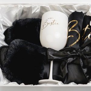 Könnte beinhalten: Eine weiße Geschenkbox gefüllt mit einem schwarzen Satin-Morgenmantel, schwarzen flauschigen Hausschuhen und einem weißen Weinglas mit dem Text "bride's bestie" in goldenen Buchstaben.