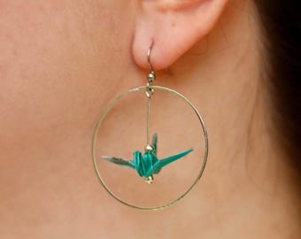 May include: A close-up of a silver hoop earring with a teal origami crane suspended inside. The crane is folded with intricate detail, and the earring is attached to a silver hook.