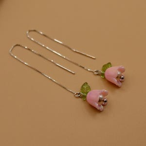 Op de afbeelding: Een paar delicate oorhangers met roze tulpvormige kralen, een zilverkleurig kralenmiddelpunt en een klein groen blad. De oorbellen hangen aan een dunne, zilverkleurige ketting.