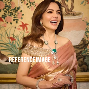 Puede incluir: Una mujer con un sari dorado y rosa con un escote dorado adornado, adornada con un collar, pendientes, pulsera y anillo de diamantes y esmeraldas. El fondo presenta una pintura floral y el texto "REFERENCE IMAGE".