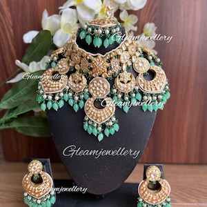 Puede incluir: Un conjunto de collar y pendientes de novia indio en oro y verde con un gran colgante. El collar presenta diseños intrincados y cuentas verdes. Los pendientes también están decorados con cuentas verdes y tienen un diseño similar al colgante.