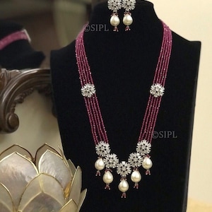 May include: A necklace and earring set on a black display. The necklace has multiple strands of small, red beads, with floral-shaped, clear crystal accents and large, pear-shaped white pearls. The earrings match the floral and pearl design.