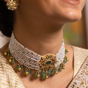 May include: A multi-strand pearl choker necklace with a gold pendant featuring a large oval green gemstone and smaller red gemstones. The necklace has dangling green beads and gold accents. Matching gold flower earrings are also visible.