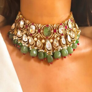 May include: A gold choker necklace featuring large, clear, faceted stones, smaller red stones, and green stones. The necklace has a layered design with dangling green and red stones.