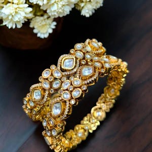 Pode incluir: Duas pulseiras douradas adornadas com pedras preciosas transparentes. As pulseiras apresentam um design intrincado com um padrão repetitivo de pedras redondas e quadradas, cravejadas em ouro. Flores brancas num vaso de madeira ao fundo.