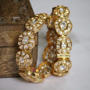 Op de afbeelding: Twee gouden armbanden met ingewikkelde ontwerpen met ronde, heldere edelstenen. De armbanden zijn op elkaar gestapeld.