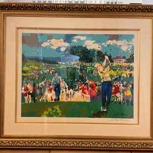 May include: A framed print of a golf scene with a golfer in mid-swing, wearing a yellow and black striped shirt and blue pants. The artwork has a colorful, impressionistic style, with a crowd of spectators and a building in the background. The frame is gold.