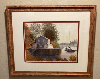 Puede incluir: Pintura a la acuarela de una escena costera con un edificio en un muelle y un velero. La obra de arte está enmarcada en un marco de madera marrón rústico. La pintura presenta una paleta de colores apagados con tonos de azul, marrón y verde.