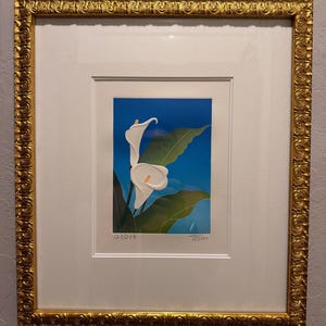 May include: A framed print featuring two white calla lilies with yellow spadices against a blue background and green leaves. The artwork is numbered 128/500 and signed by the artist. It is enclosed in an ornate, textured gold frame.