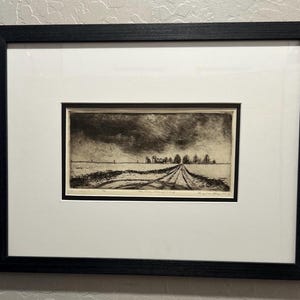 May include: A framed black and white landscape print. The artwork depicts a rural scene with a road leading towards a distant horizon under a cloudy sky. The print is matted in white and framed in a black wooden frame.
