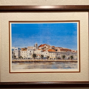 May include: A framed print of a watercolor painting depicting a cityscape with white buildings and red tile roofs. The buildings are clustered together along a waterfront with a blue sky and water in the background. The print is signed "Lisbon Harbor" by the artist.