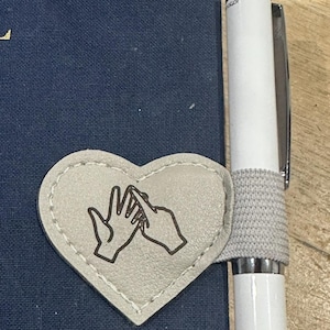 May include: A heart-shaped, light grey leather bookmark with a stitched border and a graphic of two hands signing. The bookmark rests on a navy blue notebook next to a white pen with a grey elastic band.