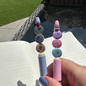 May include: Two pens, one pale blue and one pink, each with a series of small, round, decorative elements. The elements feature hand-drawn images. The pens are held over an open notebook.