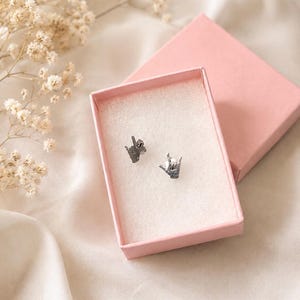 Pu&ograve; includere: Un paio di orecchini a lobo color argento a forma di mani che formano il segno della lingua dei segni americana per ti amo. Gli orecchini sono esposti all'interno di un piccolo portagioie rettangolare in cartone rosa chiaro con coperchio.