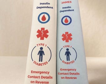 Puede incluir: Dos fundas para cinturón de seguridad, una crema y otra azul claro, con el nombre JAMES, el texto Insulin Dependent, un icono de gota de sangre roja, una estrella médica roja y un símbolo de Type 1 Diabetes. El texto Emergency Contact Details on Reverse está impreso.