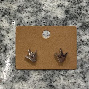 Pu&ograve; includere: Un paio di orecchini a lobo color argento a forma di mano che fa il segno ti amo nella lingua dei segni americana. Gli orecchini sono fissati su un cartoncino rettangolare marrone con un foro circolare nella parte superiore centrale.