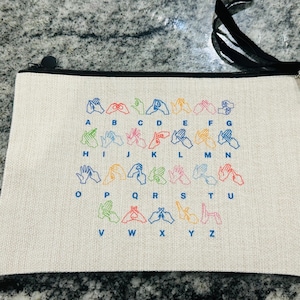 May include: Cream-coloured zippered pouch with a black zip and strap. The pouch features a colourful embroidered alphabet in British Sign Language. The letters A to Z are displayed with corresponding hand gestures.