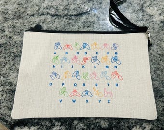 Può includere: Pochette con cerniera color crema con cerniera e cinturino neri. La pochette presenta un alfabeto ricamato colorato nella lingua dei segni americana. Le lettere dalla A alla Z sono visualizzate con i corrispondenti gesti delle mani.