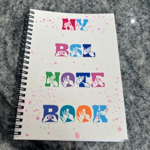 May include: A spiral-bound notebook with the text "MY BSL NOTE BOOK" in various colours. The letters are formed by hand gestures. The notebook cover is white with pink splatters.