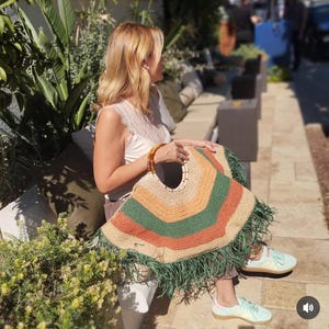 May include: A colorful, handmade tote bag with a circular handle. The bag features a geometric design with orange, green, and tan stripes, and a fringe trim. The bag is held by a person wearing a white top and light blue sneakers.