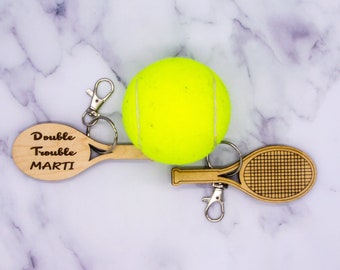 May include: A wooden tennis racket keychain with the text "Double Trouble MARTI" and a yellow tennis ball keychain. The keychains are on a white marble surface.